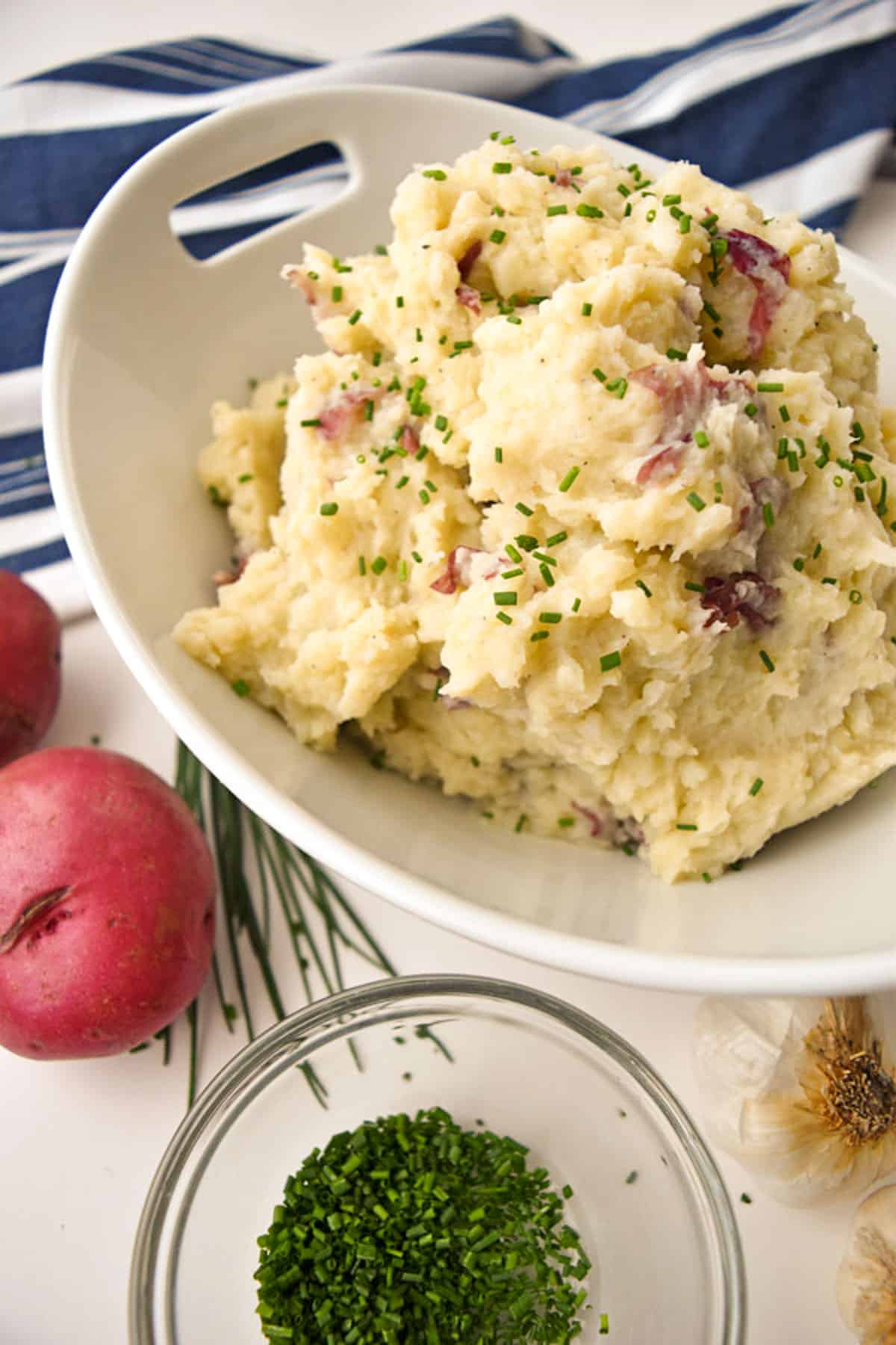 a bowl of low FODMAP mashed potatoes next to a blue and white striped towel, red potatoes and chives