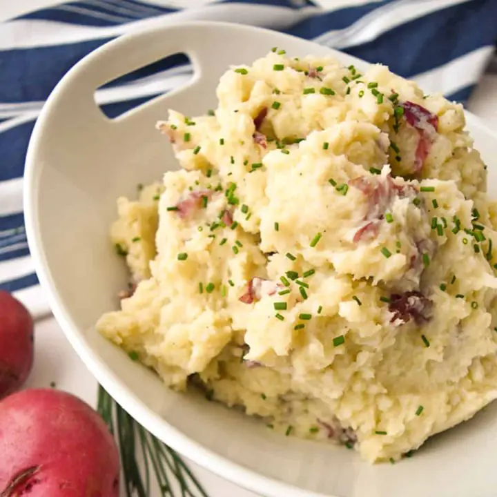 mashed potatoes in a white bowl next to red potatoes, chives, and a blue and white striped towel.