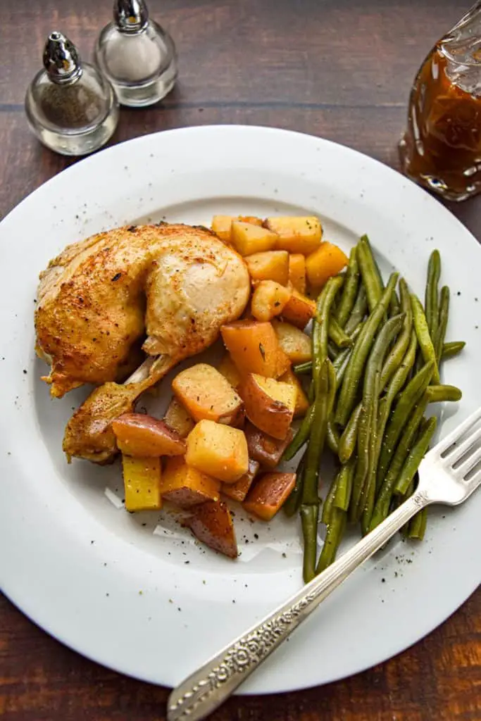 chicken thigh, potatoes and green beans on a white plate with a fork next to salt and pepper shakers and a pitcher of chicken drippings on a brown wood background