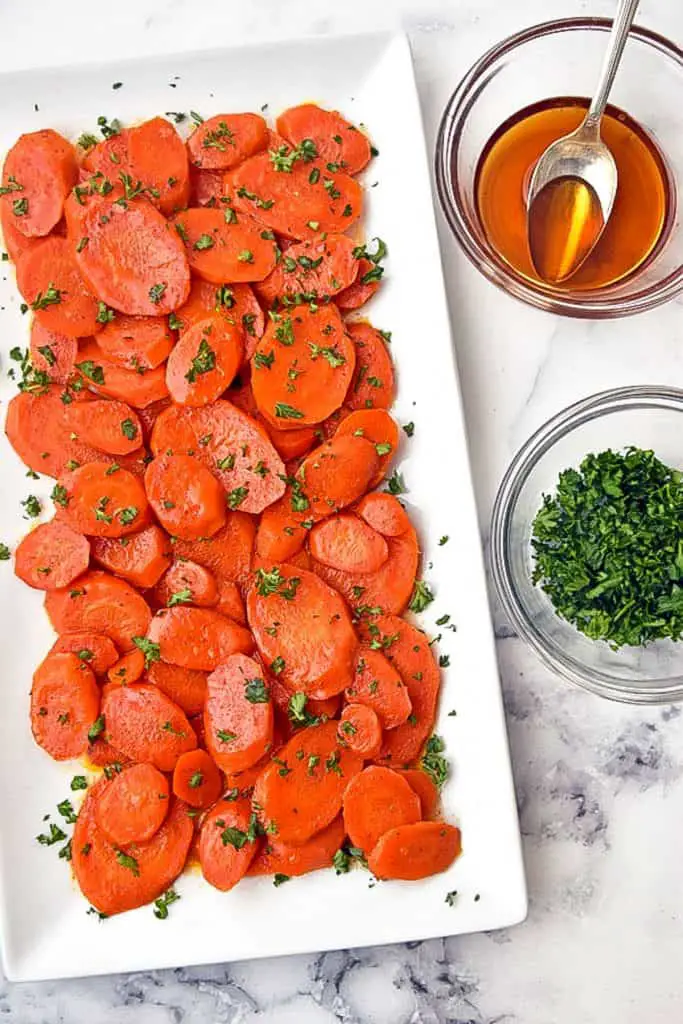 low fodmap Instant Pot Glazed Carrots on a white plate in front of bowls of maple syrup and chopped parsley
