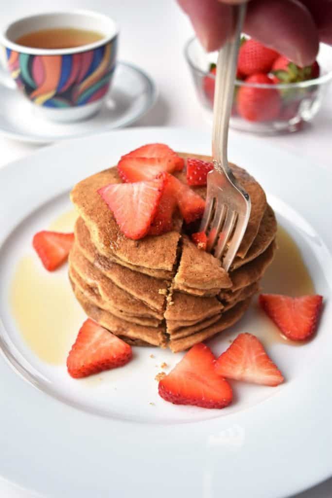 A fork taking a slice out of a stack of tigernut flour pancakes topped with strawberries and maple syrup.