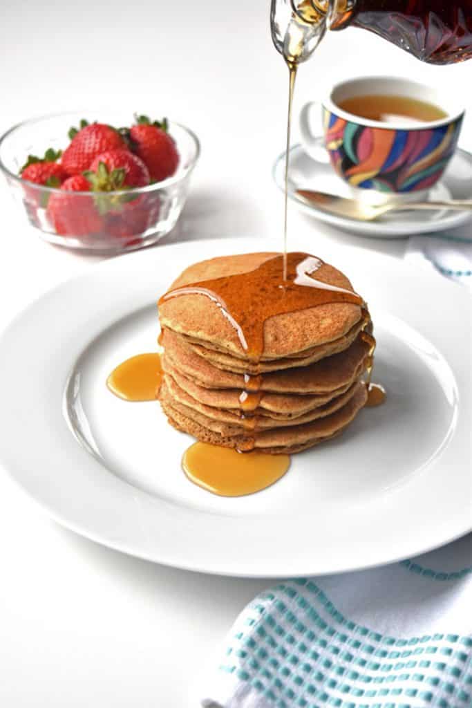 Pouring maple syrup on tigernut flour pancakes on a white plate in front of a bowl of strawberries and a cup of tea. 