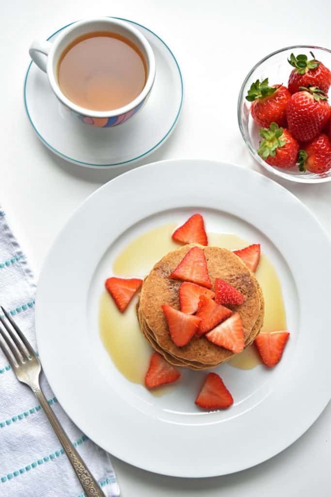 An overhead shot of a plate of stacked tigernut flour pancakes topped with strawberries and maple syrup next to a bowl of strawberries and a cup of tea
