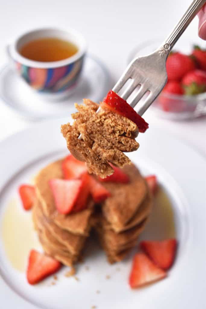 a fork holding a bite of tigernut flour pancakes topped with a strawberry in front of a stack of pancakes on a plate.