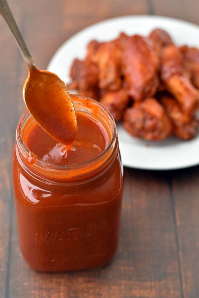 dipping a spoon in a jar of low fodmap BBQ sauce in front of a plate of BBQ chicken wings