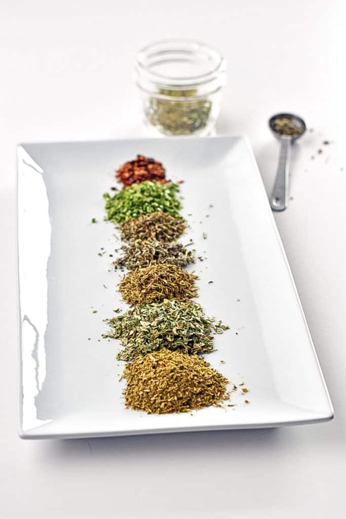 A white rectangular plate with piles of individual spices with a jar and teaspoon behind it containing low fodmap Italian seasoning