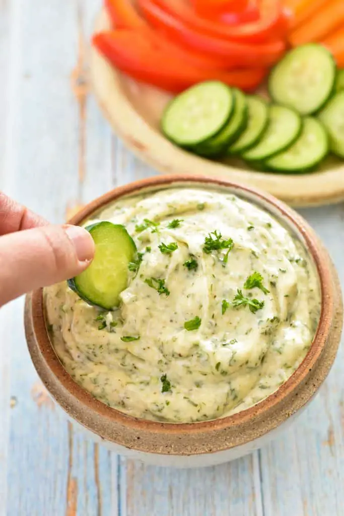 Dipping a slice of cucumber in a bowl of low fodmap veggie dip (dairy-free, paleo, whole30) that's in front of a plate of chopped veggies