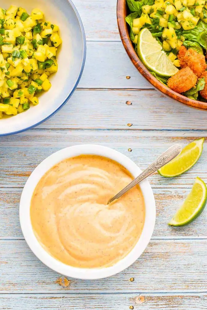 a bowl of low FODMAP chipotle mayo with a spoon inside next to lime wedges and in front of a bowl of pineapple salsa and an air fryer shrimp taco bowl