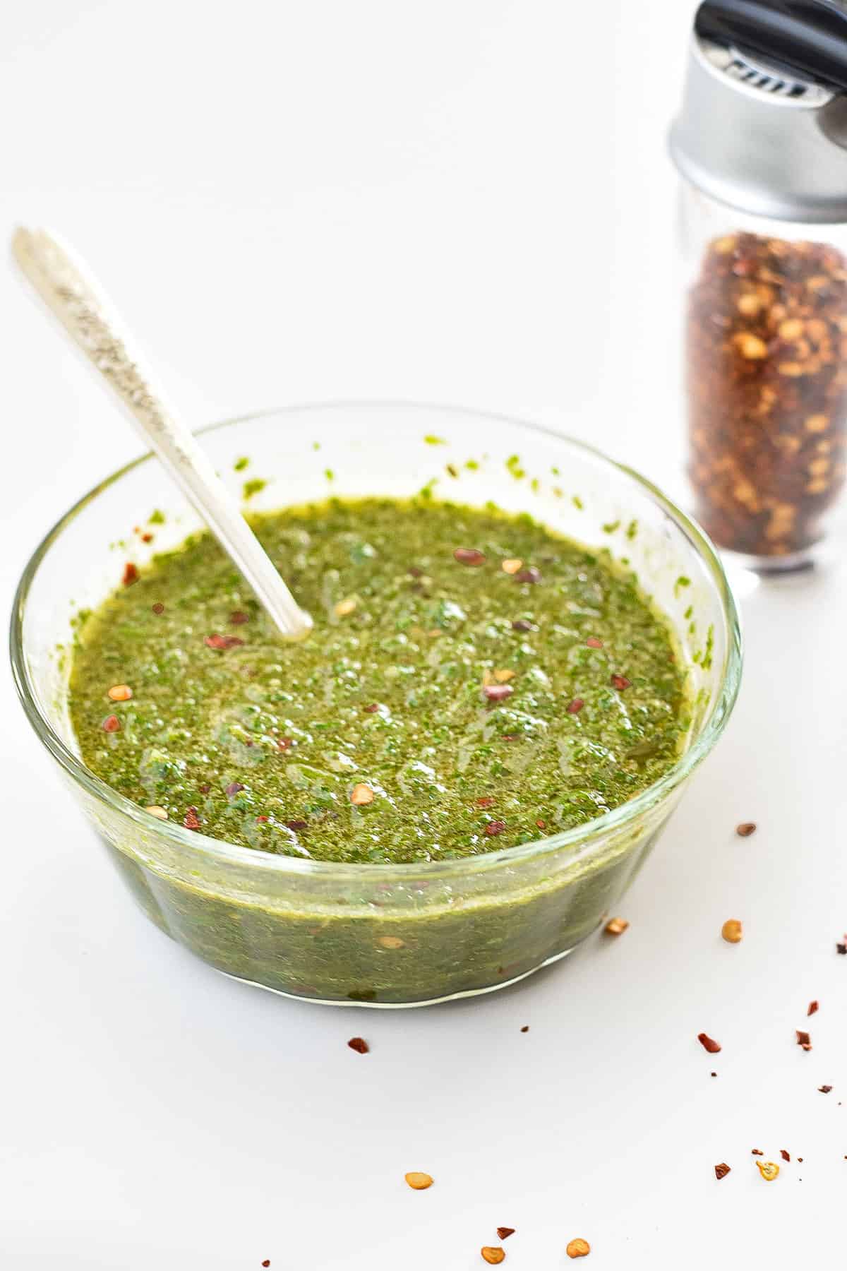 a bowl of low fodmap chimichurri sauce with a spoon in front of a shaker of red pepper flakes