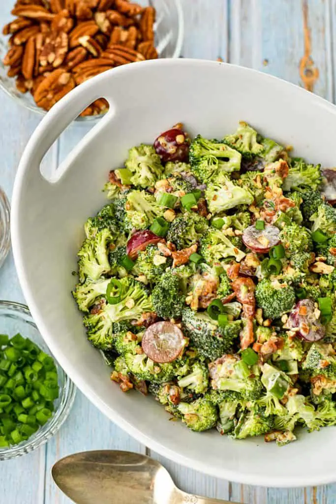 overhead shot of a bowl of low broccoli salad with bacon next to bowls of chopped green onions and pecans, and a spoon