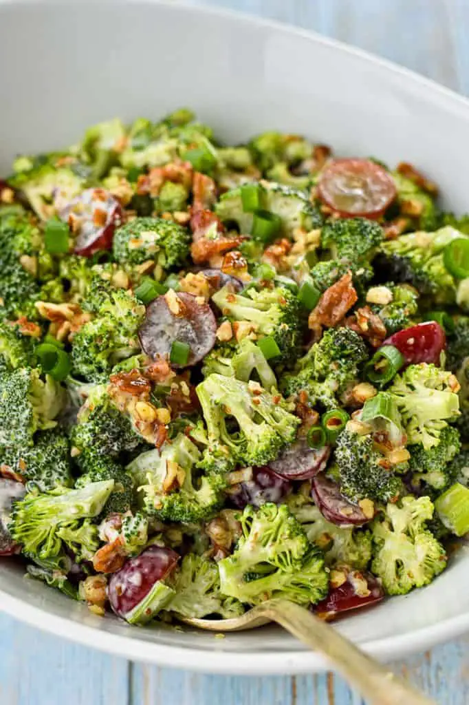 a close up of a white bowl of broccoli salad with bacon and a spoon