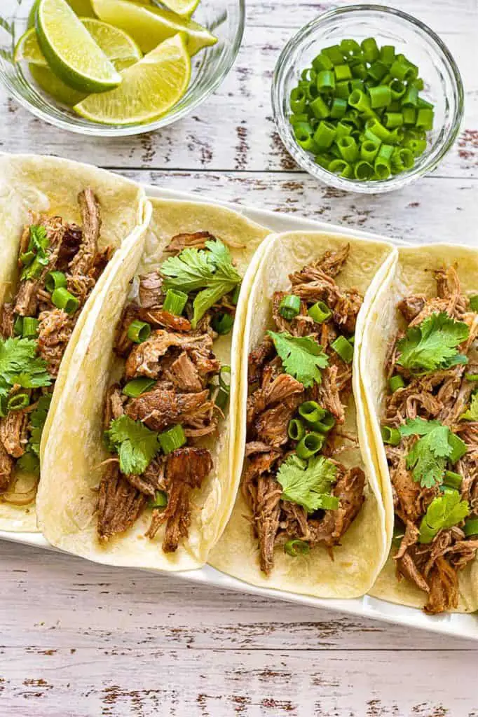 low fodmap carnitas in tortillas on a plate next to small bowls of lime wedges and chopped scallions