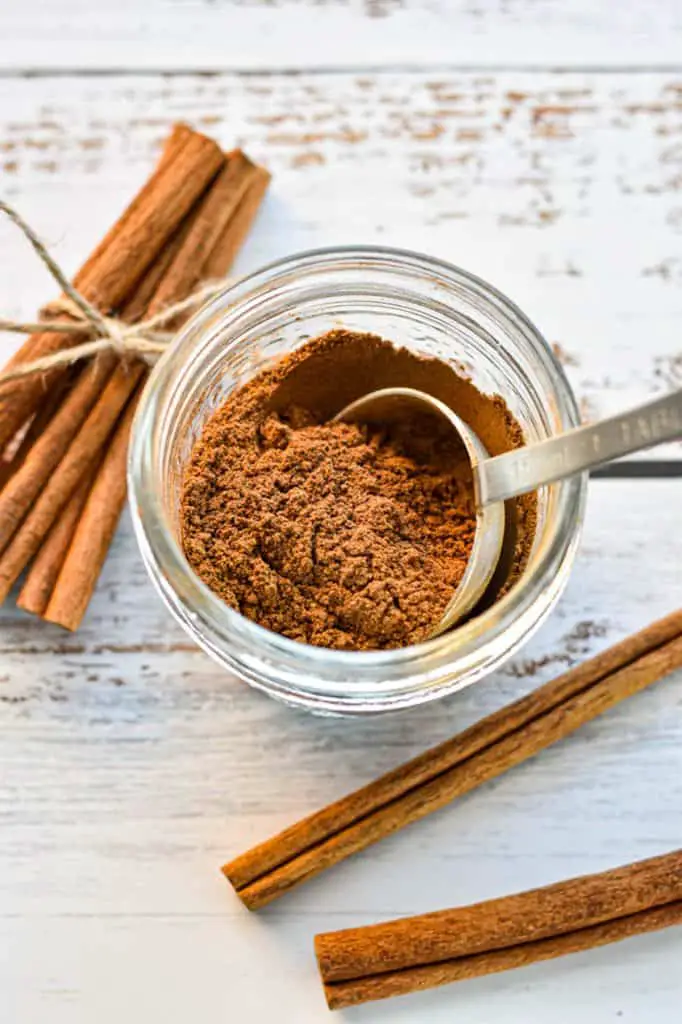 a spoon scooping a tablespoon of low fodmap homemade pumpkin pie spice out of a jar that's next to cinnamon sticks