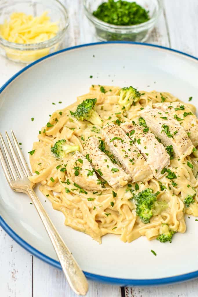 low fodmap chicken fettuccine alfredo with broccoli on a blue rimmed white plate with a fork