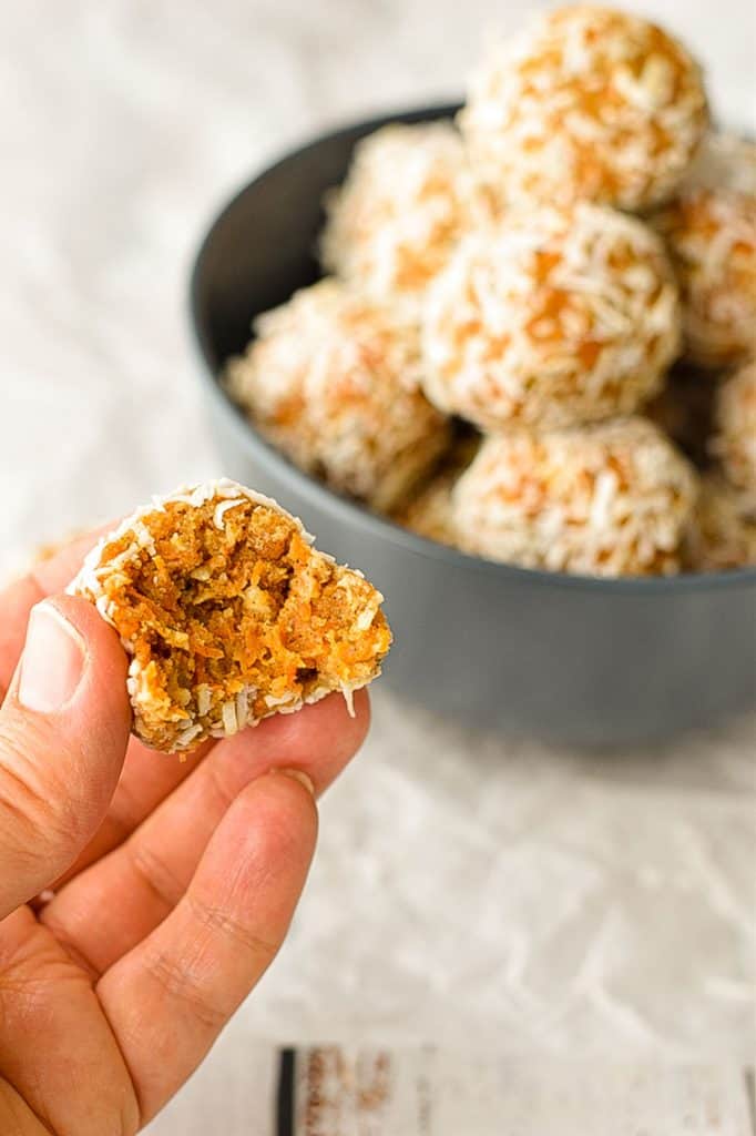 a hand holding a bitten low fodmap carrot cake energy ball with a bowl of energy balls in the background