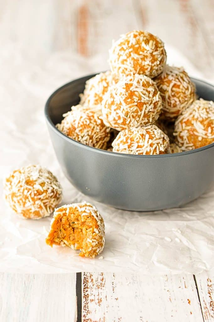 a navy blue bowl of low fodmap carrot cake bliss balls on a piece of wrinkled parchment paper next two two balls, one that has been partially eaten