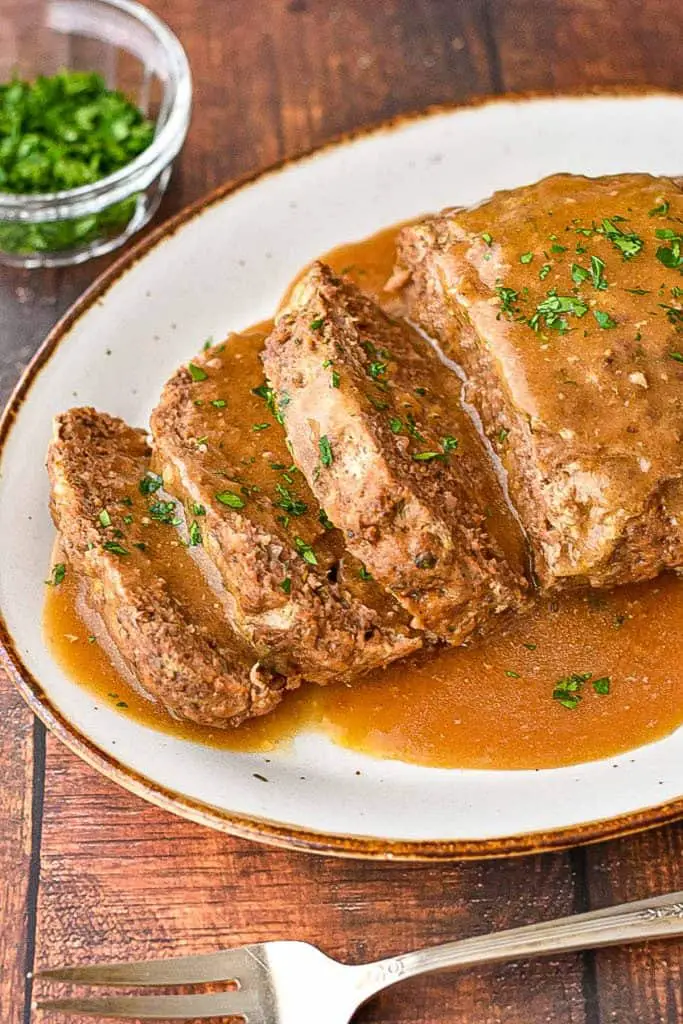 partially sliced low fodmap meatloaf on a brown rimmed platter covered in gravy and garnished with Italian parsley