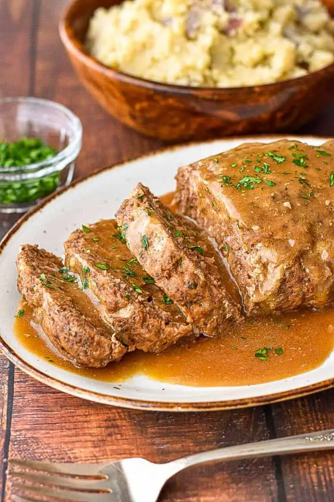 sliced low fodmap instant pot meatloaf covered in gravy in front of a wooden bowl of mashed potatoes and a glass bowl of chopped parsley