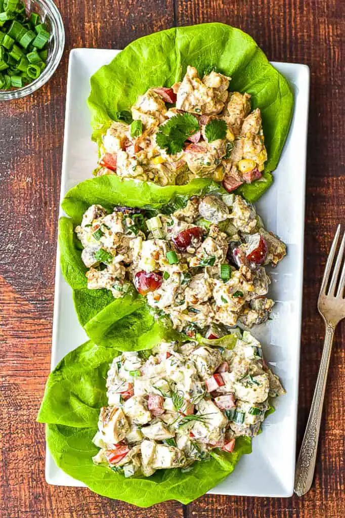low fodmap chicken salad - southwest, classic, and Mediterranean - cupped in butter lettuce on a white, rectangular plate next to a fork and a bowl of chopped scallions on a brown wood background