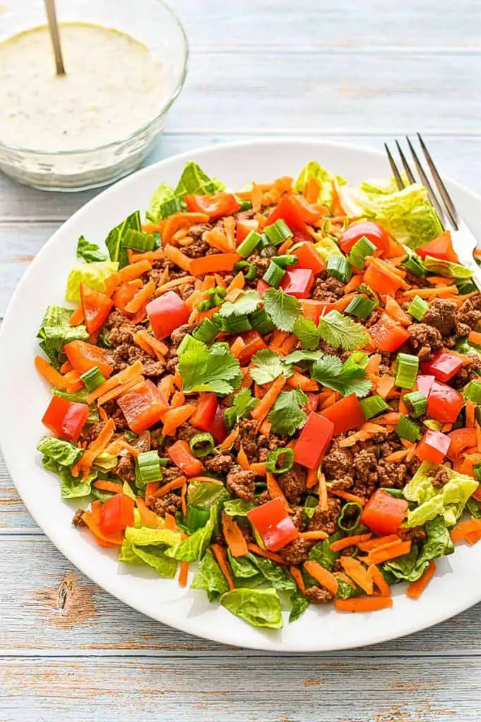 a low fodmap taco salad with ground beef lettuce, red bell pepper, green onion, carrots, and cilantro on a white, round plate in front of a bowl of ranch dressing on a blue background