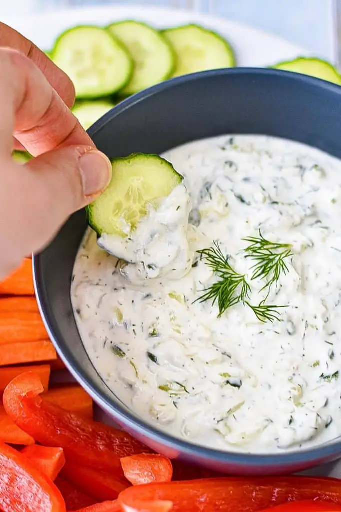 a cucumber being dipped into a navy blue bowl of low FODMAP tzatziki on a plate of chopped vegetables