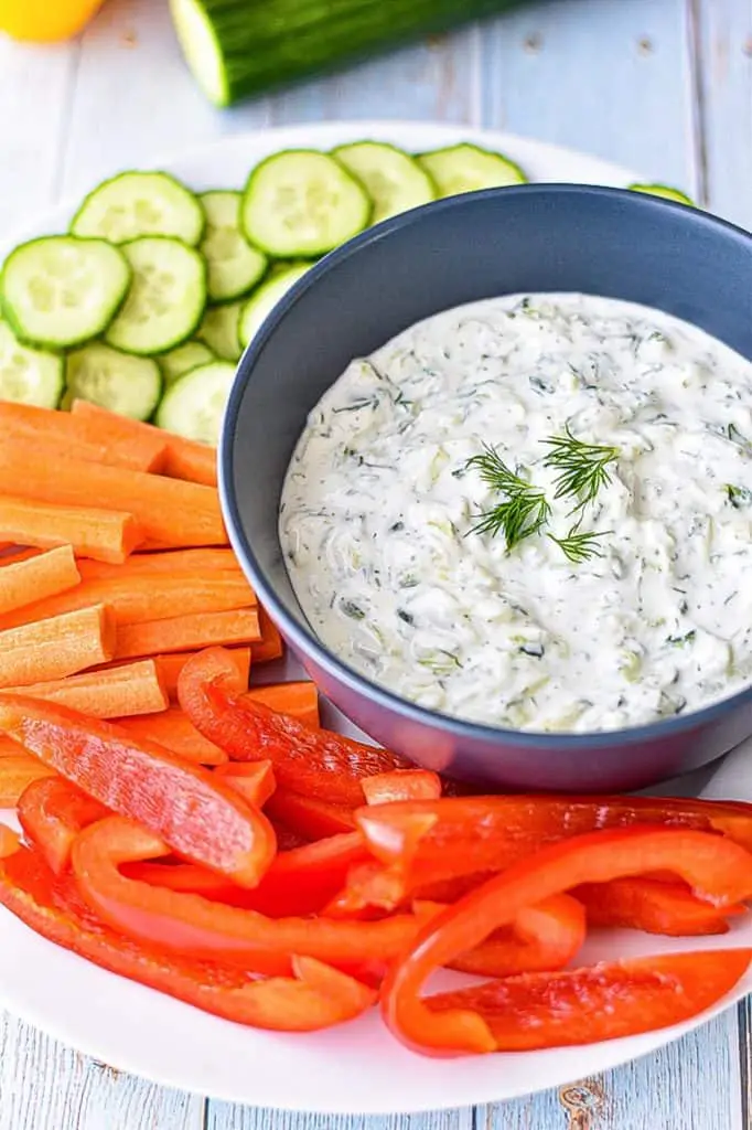 a blue bowl of low FODMAP tzatziki sauce on a plate of veggies and garnished with fresh dill
