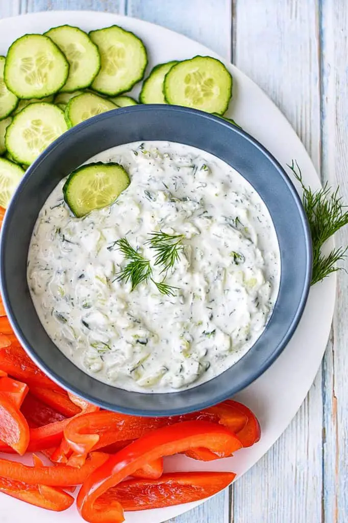 low fodmap tzatziki in a navy blue bowl on a white plate covered in chopped veggies and garnished with fresh dill leaves