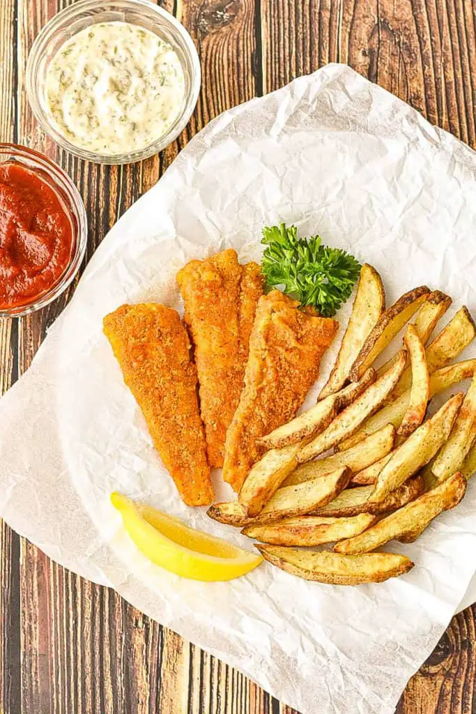 overhead shot of gluten-free fish and chips on a parchment paper covered plate with lemon and parsley next to bowls of ketchup and tartar sauce