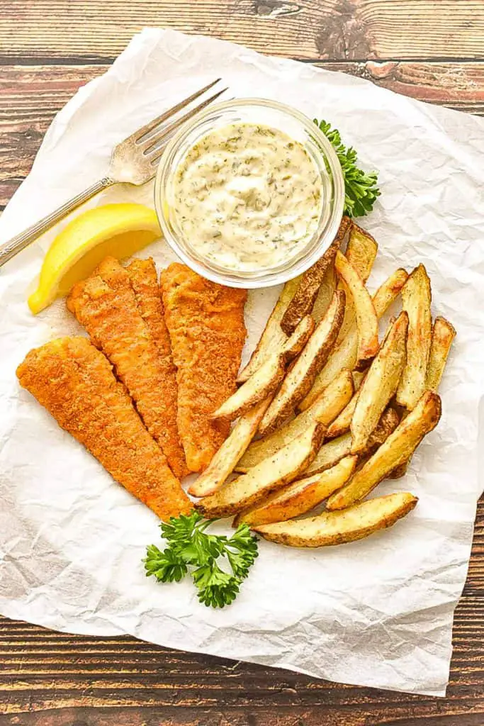 low fodmap fish and chips made with the air fryer on a plate covered in parchment paper with a bowl of tartar sauce, slice of lemon, sprigs of parsley, and a fork