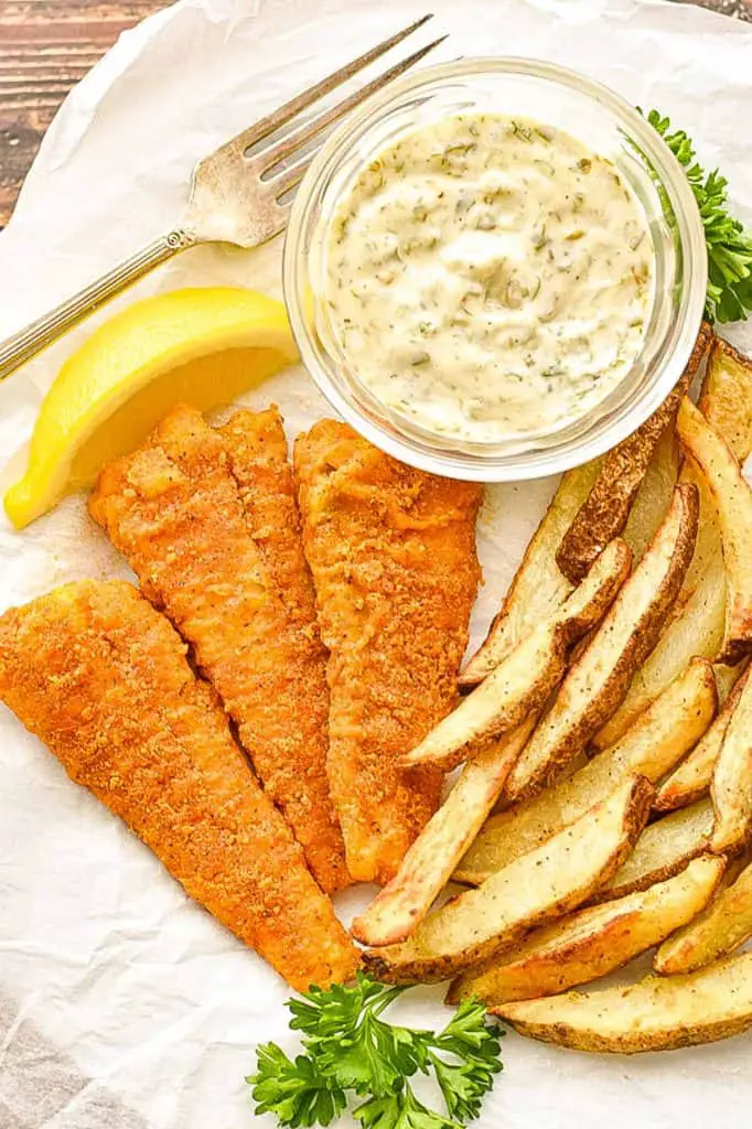 close up shot of low fodmap fish and chips, garnished by a slice of lemon and parsley springs on white parchment paper with a fork
