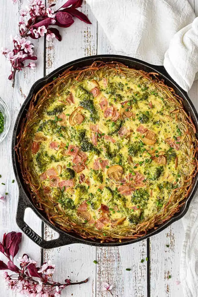 low fodmap quiche with a gluten-free hash brown crust in a cast iron skillet next to pink flowers on a white background