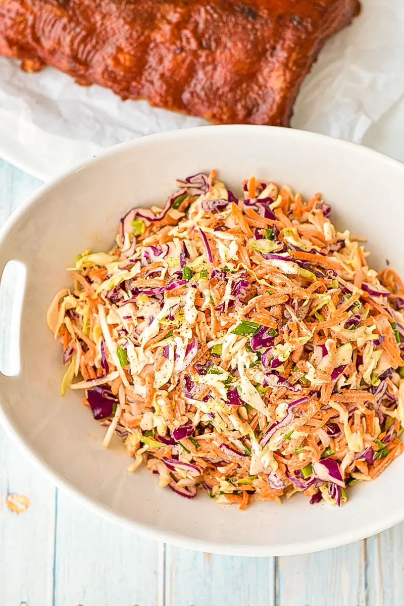overhead shot of low fodmap coleslaw in a white bowl with handles in front of a rack of barbecue ribs on a light blue background