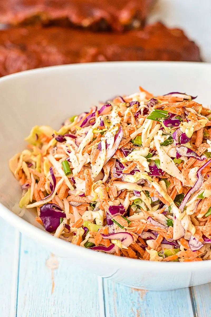 a white bowl of low fodmap coleslaw in front of barbecue ribs on a blue background