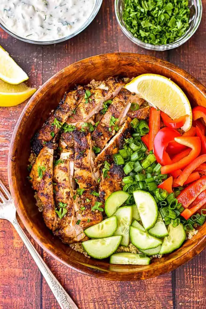low FODMAP grilled chicken shawarma in a wooden bowl with sliced red bell pepper, cucumber, scallions, and quinoa next to bowls of tzatziki and chopped parsley and a few lemon slices