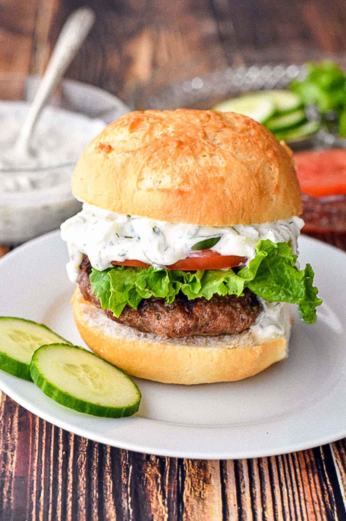 low fodmap gyro burger topped with tzatziki, cucumber and tomato on a low fodmap hamburger bun next to sliced cucumber on a white plate