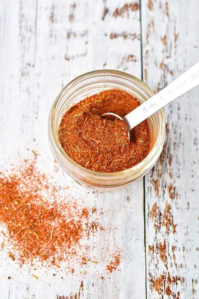 overhead shot of low fodmap creole seasoning mix in a clear jar with a teaspoon on a wooden background painted white