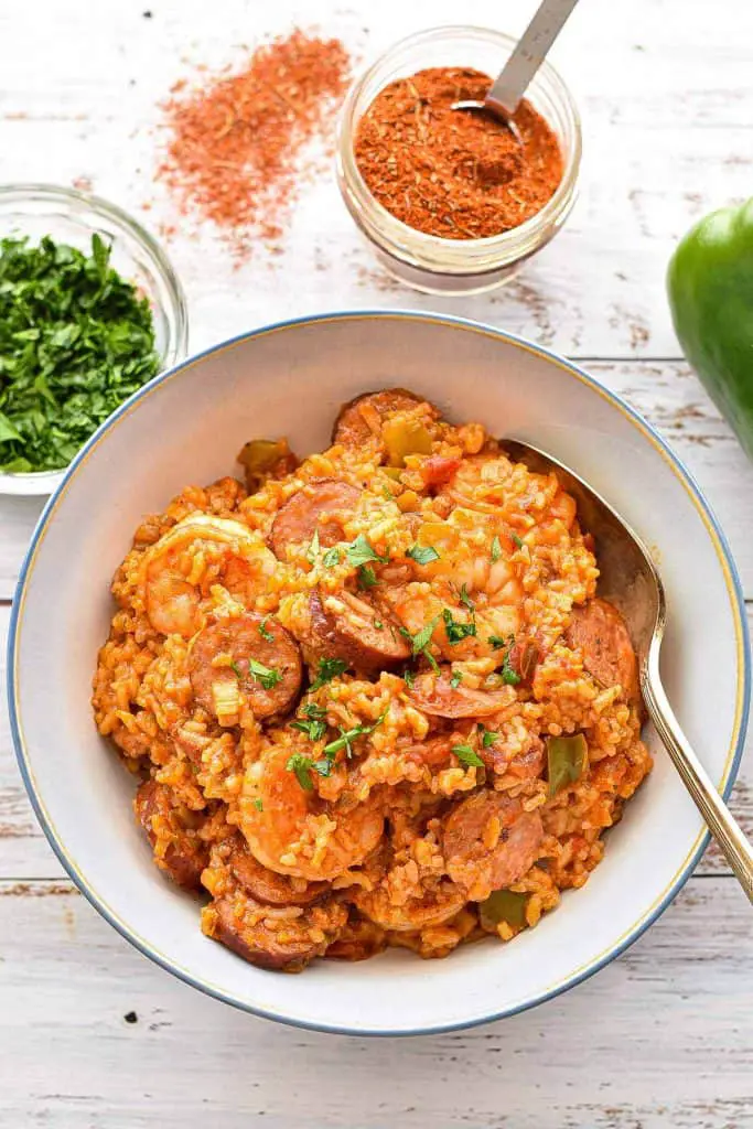 overhead shot of low fodmap jambalaya in a blue rimmed bowl with a spoon next to a bowl of chopped parsley, creole seasoning and a green bell pepper