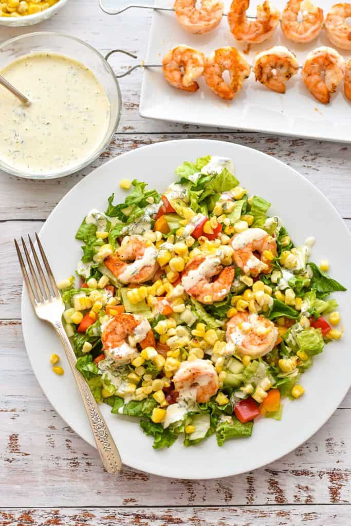 overhead shot of low fodmap detox salad with shrimp and corn dressed in dairy-free low fodmap ranch dressing on a white plate with a fork in front of a plate of shrimp skewers and a bowl of ranch dressing