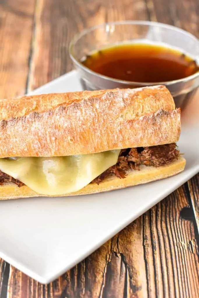 a french baguette with beef and cheese  on a white rectangular plate with a clear bowl of au jus