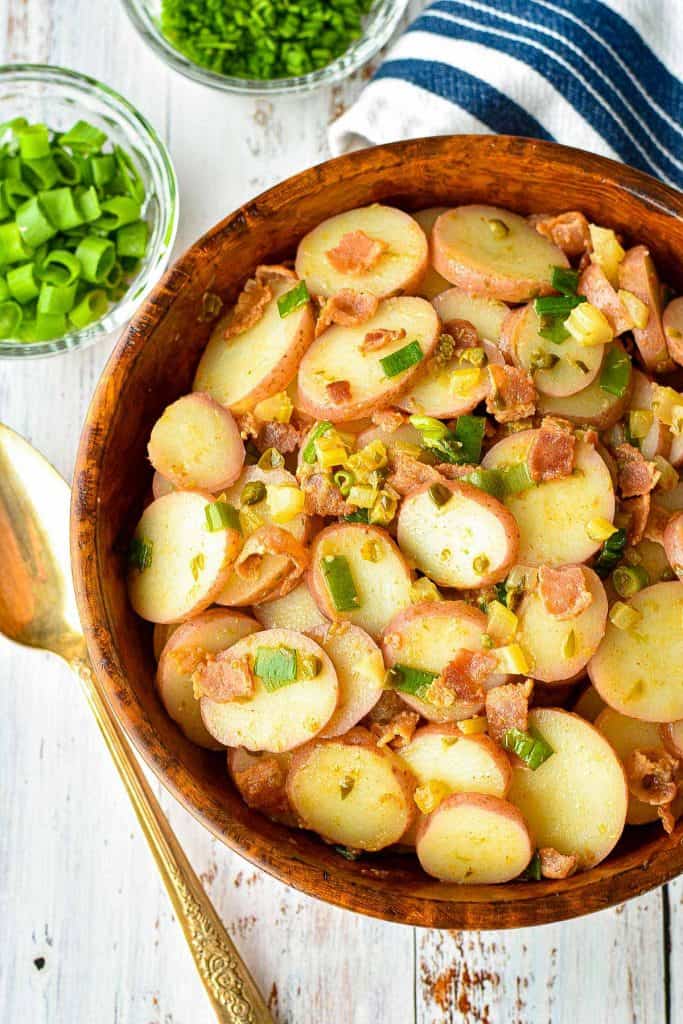 low fodmap instant pot kartoffelsalat german potato salad in a wooden bowl next to a spoon, a blue and white striped towel, and bowls of chopped green onions and chives