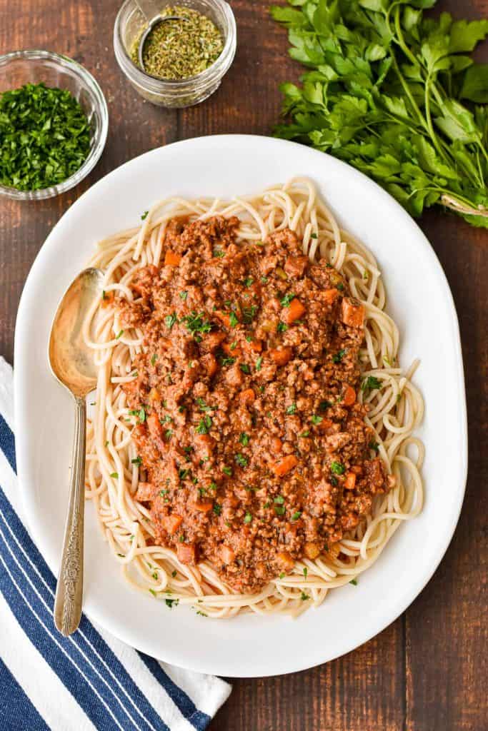 overhead shot of low fodmap Bolognese on spaghetti on a white platter with a spoon next to a bunch and bowl of parsley, jar of italian seasoning, and a blue and whit striped towel.