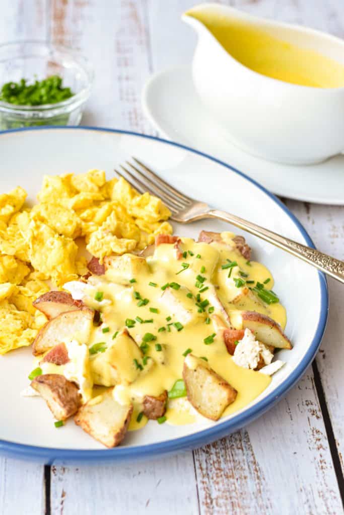a blue and white plate of low fodmap breakfast poutine and scrambled eggs next to a gravy boat of low fodmap hollandaise sauce and a bowl of chopped chives