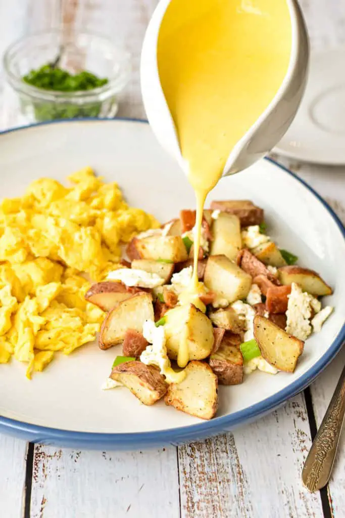pouring low fodmap hollandaise sauce on low fodmap breakfast poutine next to scrambled eggs on a blue and white plate