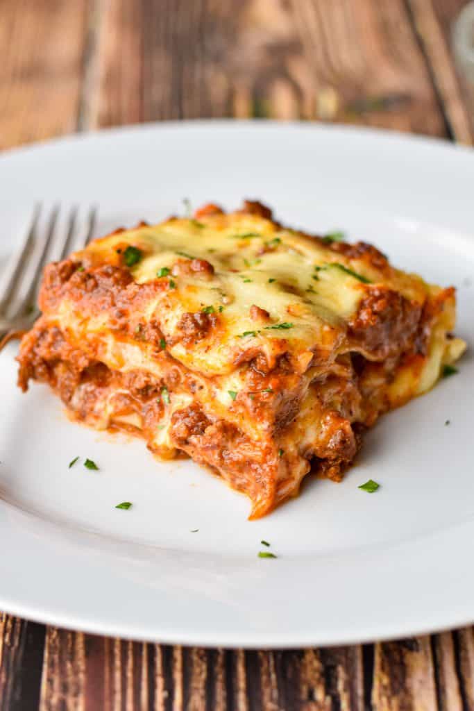 a square piece of low fodmap lasagna Bolognese on a white plate with a fork on a wooden background.