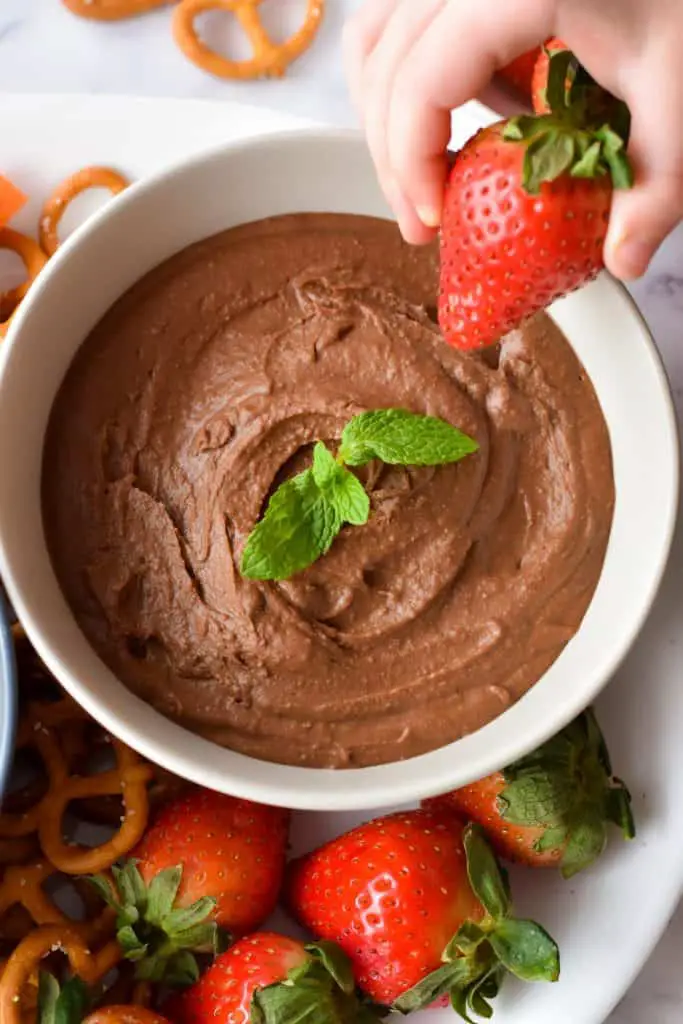 dipping a strawberry into low fodmap chocolate dessert in a white bowl on a platter with strawberries and pretzels