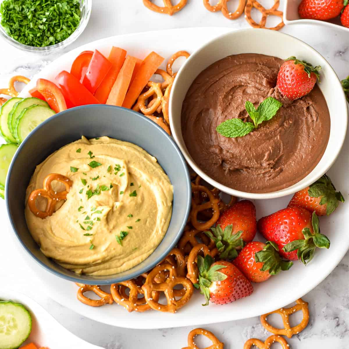 classic and dessert hummus in bowls on a platter with strawberries, pretzels, and chopped veggies