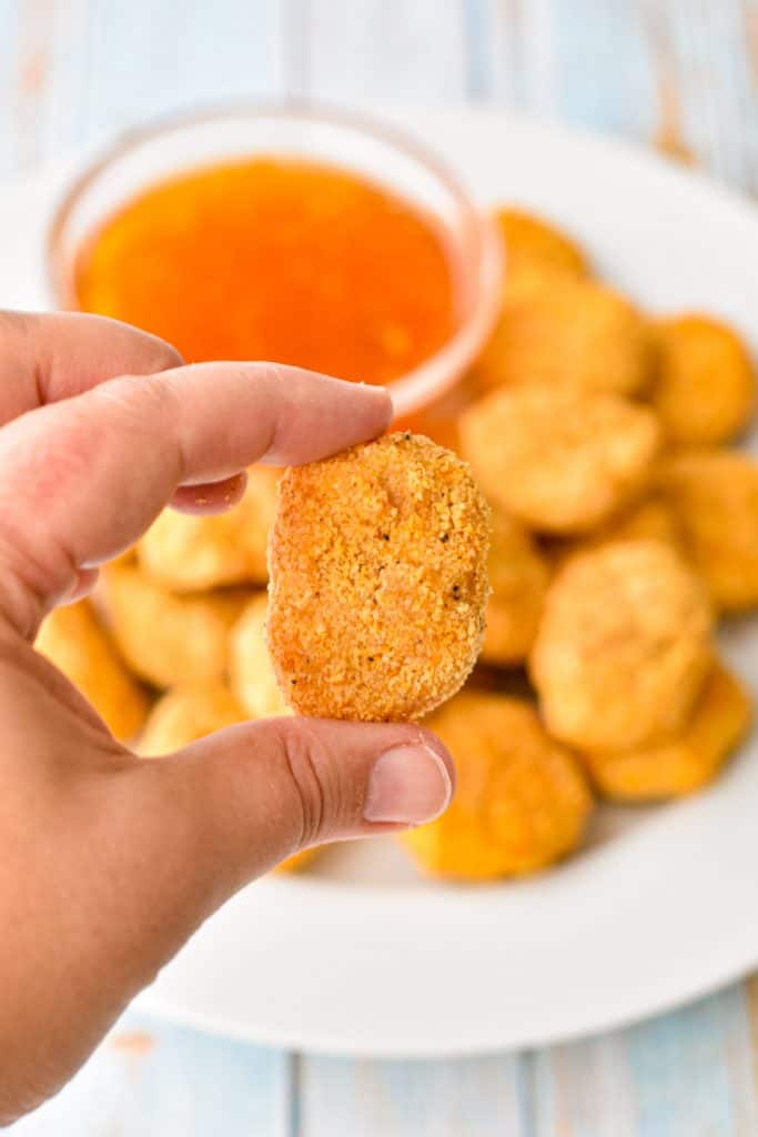 a hand holding a grain-free chicken nugget above a plate of chicken nuggets with sweet and sour sauce