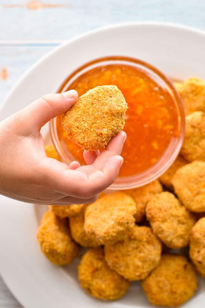 a kid's hand holding a low fodmap chicken nugget over a bowl of low FODMAP sweet and sour dipping sauce and a plate of low fodmap chicken nuggets