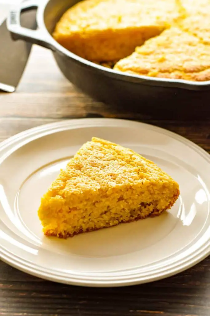 a triangular slice of low fodmap cornbread on a white plate in front of a skillet of cornbread 