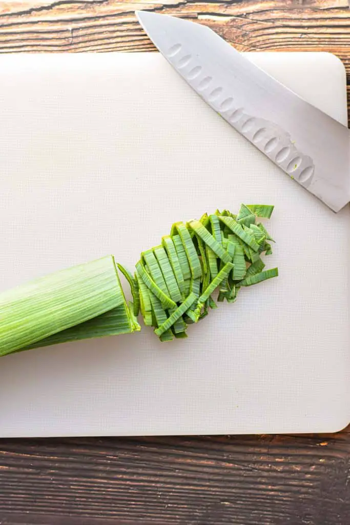 the dark green leaves of a leek sliced horizontally on a white chopping board next to a knife.